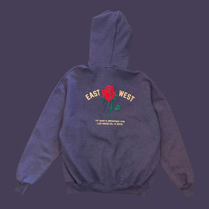 The same purpley grey zip-up hoodie from the back view on a white background. The hoodie has a red rose embroidered on the center. To the left of the rose it says “EAST” and to the right it says “WEST” under either the rose reads “ 727 NORTH BROADWAY #115 LOS ANGELES, CA 90012”.