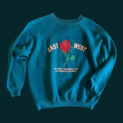A sweatshirt with embroider that reads "East West 727 N. Broadway #115, Los Angeles, CA 90012".  The sweatshirt is a dark blue green color and features raglan sleeves